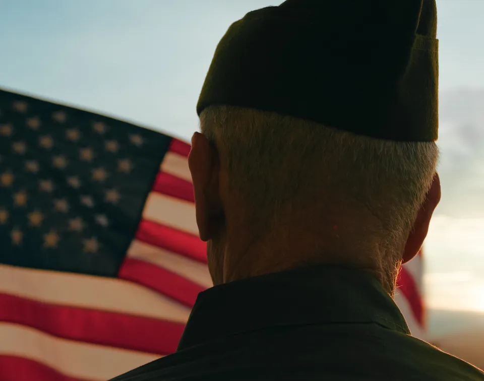 A veteran in a garrison cap, seen from behind, facing an American flag at sunset